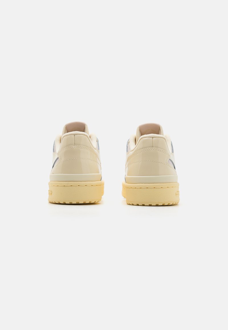 Zapatillas de piel sintética blanca con acentos en beige, puntera redonda, suela plana y parte trasera texturizada. Presentan un branding minimalista y un acabado suave.