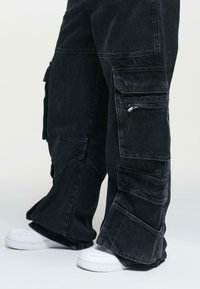 Baggy black denim cargo pants with multiple pockets and zipper details, paired with white sneakers. Fabric has a slightly textured finish.