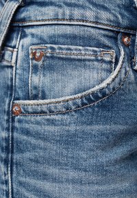 Jasnoniebieskie dżinsy mają tylną kieszeń z brązowym metalowym guzikiem oraz małym akcentem logo. Materiał ma lekko zużytą teksturę.