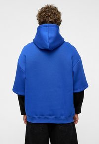 Blauwe oversized hoodie van zachte stof, met een grote capuchon, korte mouwen en een ontspannen pasvorm, gecombineerd met zwarte broek.