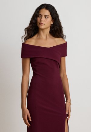 Femme aux longs cheveux foncés portant une robe bordeaux sans manches et épaules dénudées avec une fente latérale, debout devant un fond clair uni.