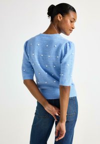Maglione azzurro chiaro lavorato a maglia con maniche a sbuffo corte, caratterizzato da pois bianchi. Texture morbida e dettagli a coste sull'orlo, abbinato a jeans scuri.