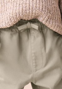 Pantaloni verde chiaro con vita elastica e coulisse, parzialmente coperti da un maglione beige a maglia con trama. Tessuto dalla finitura liscia.