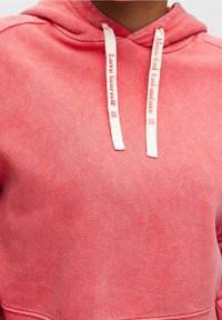 Sudadera de color coral rosa con una textura suave, que presenta un cordón con "Ámate a ti mismo" estampado en un lado y un bolsillo tipo canguro.