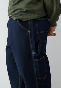 Pantalons en denim bleu marine avec des coutures jaunes visibles, dotés de poches latérales et d'une coupe décontractée. Le tissu semble robuste et texturé.