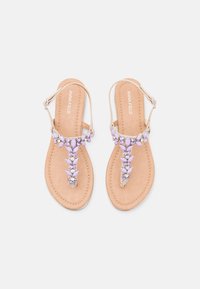 Sandales plates beiges avec un design à lanière en T, ornées d'accents de gemmes lilas et transparentes sur le dessus, avec une semelle intérieure texturée.