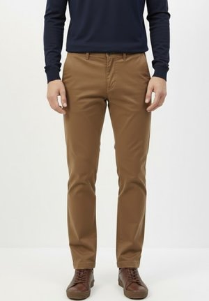 Hombre que lleva pantalones ajustados marrones, camisa de manga larga azul oscuro y zapatillas de cuero marrón, de pie frente a un fondo claro.