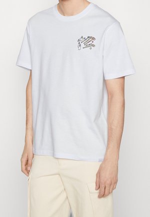 T-shirt blanc à manches courtes avec un petit graphisme sur la poitrine représentant des maillets de croquet, des balles et des bouteilles au-dessus du texte "LES DEUX", porté avec un pantalon beige.