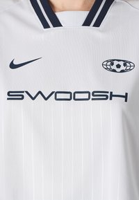 White Nike jersey with a collar, featuring navy blue accents, "SWOOSH" text, and a circular soccer graphic. Vertical textured stripes throughout.