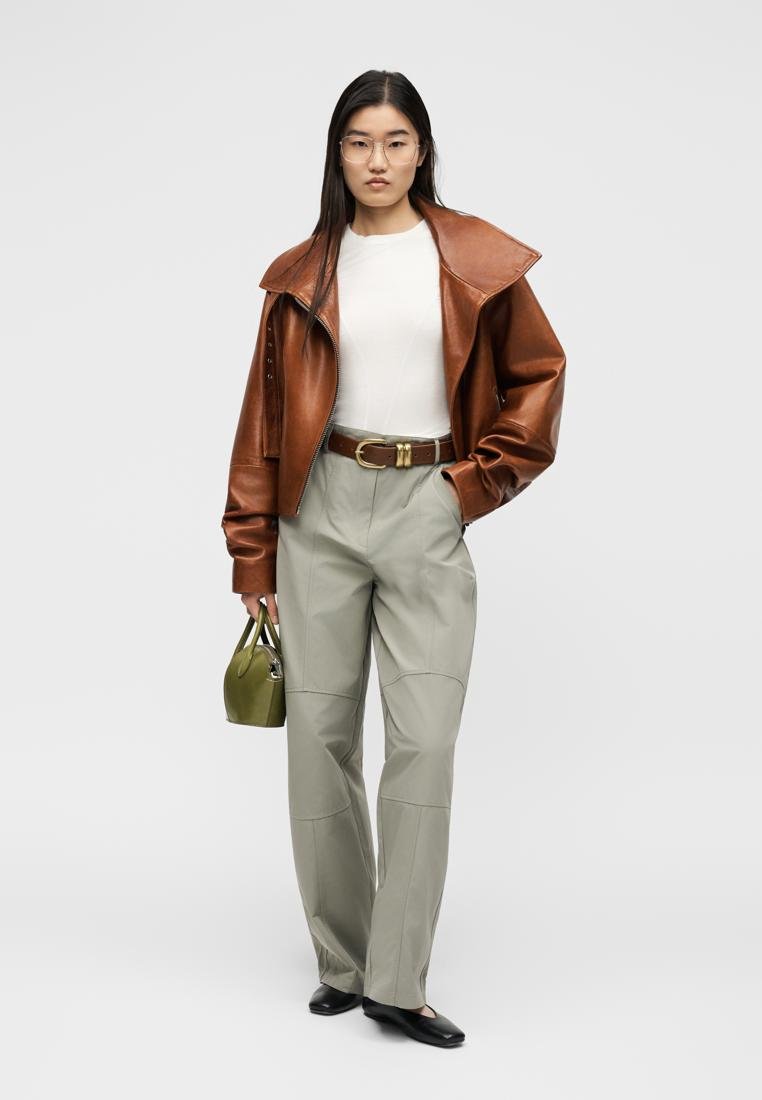 Femme portant une veste en cuir marron oversize, un haut blanc, un pantalon gris, des chaussures noires, tenant un petit sac à main vert, debout devant un fond uni.