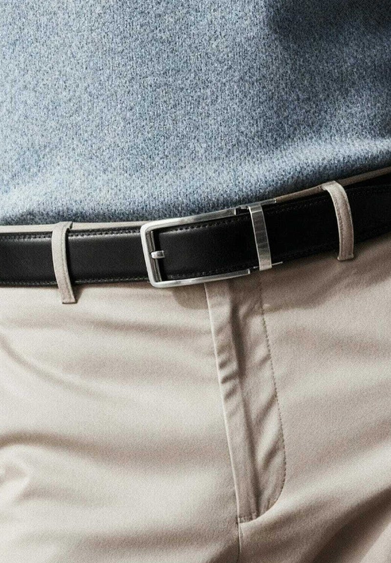 Cinture en cuir noir avec une boucle en métal argenté, portée par-dessus un pantalon beige, associée à un pull bleu clair. Texture lisse, design simple.