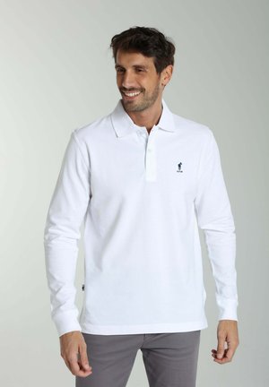 Homme souriant, portant un polo blanc à manches longues avec un petit logo sur la poitrine et un pantalon gris, debout devant un fond clair uni.