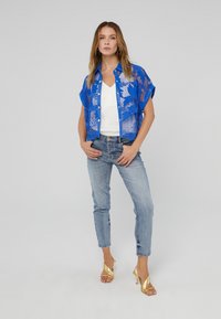 Blue patterned sheer shirt with short sleeves over a white top, paired with light wash denim jeans and gold strappy high heels.