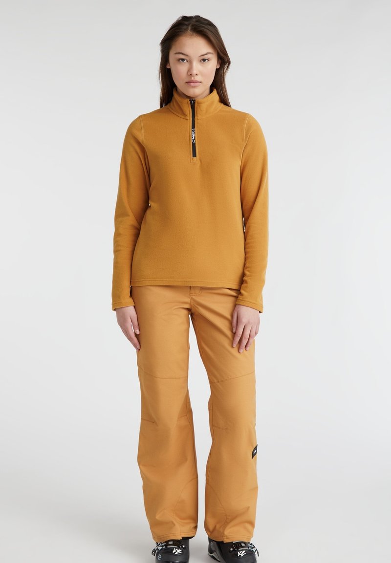 Długi rękaw, polarowa bluzka na zamek w musztardowym kolorze połączona z dopasowanymi lekkimi spodniami. Obie części mają miękką teksturę i minimalne akcenty.