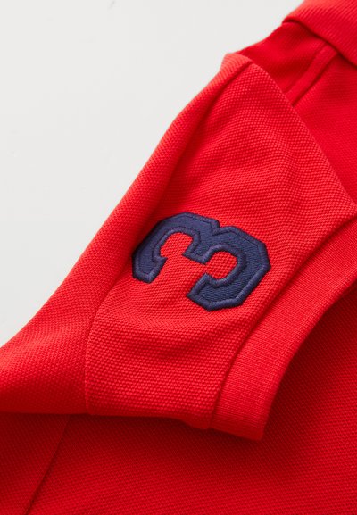 Sarkans polo krekls ar strukturētu audumu, īsām piedurknēm un jūras zila izšūta logo kreisajā plecā.