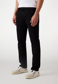 Mustat farkut slim fit -mallilla, viisi taskua ja hienovaraiset ompeluyksityiskohdat, yhdistettynä valkoisiin tennareihin, joissa on beigejä yksityiskohtia.