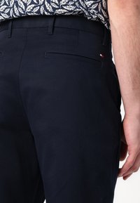 Marineblauwe pantalon van gladde stof, met een achterzak en een klein accent in rood, wit en blauw dicht bij de tailleband.