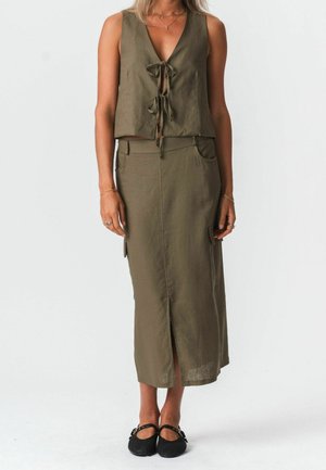 Vrouw draagt een olijfgroen mouwloos topje met voorstrikjes en een bijpassende midi-rok met hoge taille, voorsplit en zakken, gecombineerd met zwarte platte schoenen.