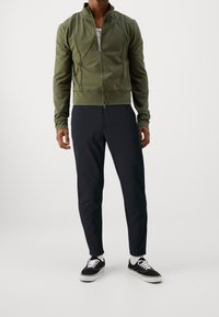 Olijfgroene zip-up jas met getextureerde naden, gecombineerd met donkere slim-fit broek en zwarte sneakers met witte zolen. Eenvoudig, modern ontwerp.