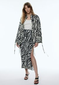 Zebra-print blouse en rok set in zwart en wit, met een losse pasvorm, lange mouwen en zijsplitten; gedragen met zwarte slide sandalen.