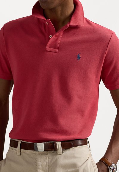 Κόκκινη πόλο μπλούζα με υφασμάτινη υφή, πλακέτα με δύο κουμπιά και μπλε λογότυπο στο αριστερό στήθος. Φορεμένη με χακί παντελόνι και καφέ ζώνη.