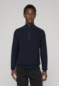 Pull-over à demi-fermeture éclair bleu marine en tissu texturé, avec un col montant et un ourlet côtelé. Le modèle le porte avec un pantalon noir, sur un fond neutre.