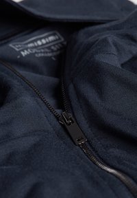 Chaqueta azul marino con cremallera y tela texturizada. Presenta una cremallera negra y una etiqueta visible en el interior del cuello, enfatizando líneas limpias y simplicidad.