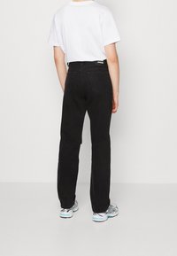 Černé džíny z denimového materiálu se standardním střihem, obsahující klasické zadní kapsy a malou černou nášivku na pase. Nosič má na sobě bílé tričko.