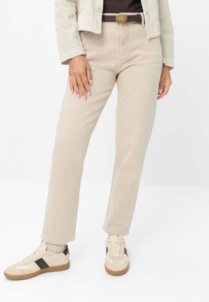 Person iført beige bukser med lige ben, hvide sneakers med sorte striber, et brunt bælte og en cremefarvet jakke over en mørk top.