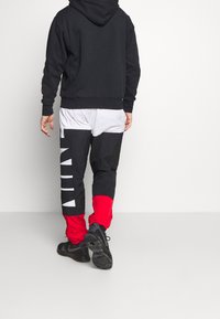 Mann trägt einen schwarzen Hoodie, farbige Hosen in Schwarz, Weiß und Rot mit großen weißen Buchstaben und schwarze Sneakers und läuft auf einem weißen Boden.