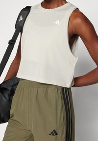 Jasnobeżowy krótki top na ramiączkach z okrągłym dekoltem i szerokimi wycięciami na ramiona. Na piersi znajduje się białe logo. Do kompletu dołączone są oliwkowozielone spodnie.