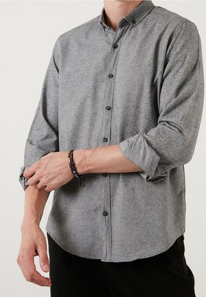 Man rolt de mouwen op van een grijs overhemd met lange mouwen en knopen, en draagt een zwartbruine kralenarmband aan zijn linker pols.