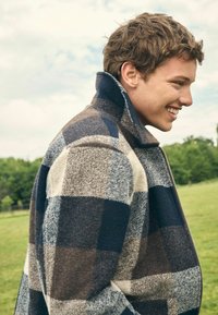 Young man smiling outdoors wearing a high-collar jacket with blue, gray, and beige large check pattern, standing on grass near trees.