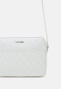 Hvid crossbody taske med Calvin Klein-logo og gentaget CK-mønster. Glat syntetisk materiale, lynlåslukning, justerbar rem.