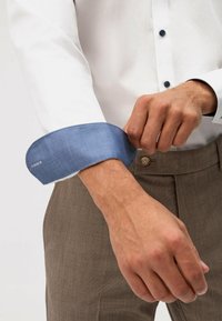 Chemise blanche avec un poignet à carreaux bleus, boutonnée jusqu'au poignet. Pantalon marron visible, avec un détail de bouton. Main ajustant le poignet.