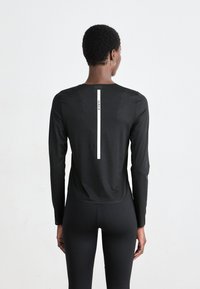Persoon met de rug naar de camera, gekleed in een zwart lange mouwen top met verticale witte streep en tekst op de rug, gecombineerd met zwarte leggings, tegen een effen achtergrond.