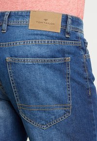 Blue denim jeans featuring a leather label with "TOM TAILOR" logo. Classic five-pocket design with visible stitching and a smooth texture.