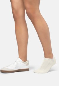 Zapatilla blanca con detalles en beige claro y lengüeta texturizada, acompañada de un calcetín de tobillo color crema. Ambos elementos están dispuestos sobre un fondo liso.