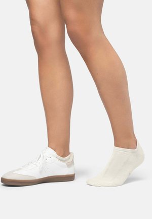 Zapatilla blanca con detalles en beige claro y lengüeta texturizada, acompañada de un calcetín de tobillo color crema. Ambos elementos están dispuestos sobre un fondo liso.