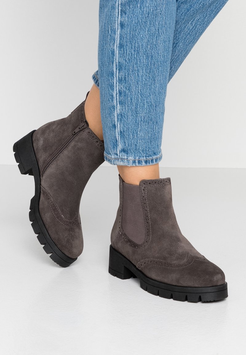 Bottes cheville en daim gris avec une surface texturée, des panneaux latéraux élastiques et une semelle noire épaisse. Détails perforés sur la zone des orteils.