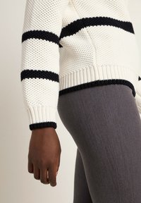 Weißer, gestrickter Pullover mit marineblauen horizontalen Streifen, mit gerippten Bündchen und Saum. Kombiniert mit grauen Leggings, die eine schlanke Passform zeigen.