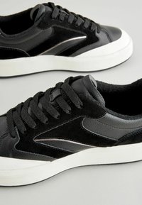 Mustad tennisud, millel on sileda naha ja sameti kombinatsioon, valge kummist tallaga ja kontrastsete mustade paeltega.
