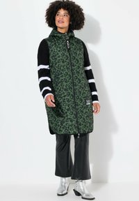 Gilet lungo in stampa leopardo verde, con chiusura a zip, cappuccio, maniche nere a righe, abbinato a pantaloni neri a campana e stivaletti argentati.