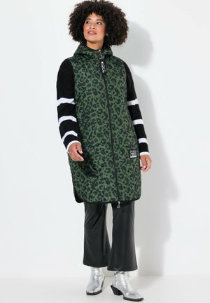 Groene luipaardprint lange vest, ritssluiting, capuchon, zwarte gestreepte mouwen, gecombineerd met zwarte flared broeken en zilveren enkellaarsjes.
