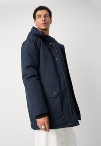 Navy blue parka jacket with a zip closure, adjustable hood, and multiple front pockets. Made from a smooth, water-resistant fabric.