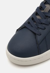 Marinblå lädersneakers med en slät yta, grå snörning och vit gummisula. Har sömdetaljer längs sidorna.
