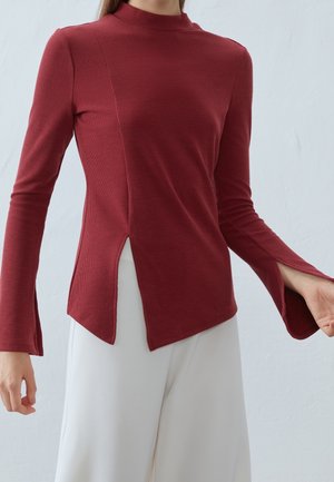 Vrouw draagt een donkerrood, geribd shirt met lange mouwen, zijslits en uitlopende manchetten, gecombineerd met witte wijde broekspijpen.