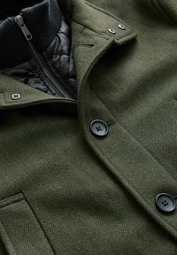 Olivgrüne Wollmischjacke mit Fischgrätenmuster, ausgestattet mit einem schwarzen gepolsterten Innenfutter, Reißverschluss und schwarzen Knöpfen.