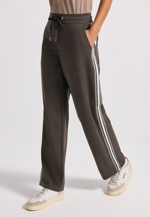 Person wearing dark brown sweatpants with white side stripes and white sneakers, standing with one hand in pocket.