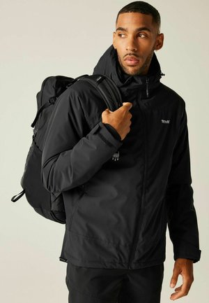 Mężczyzna w czarnej kurtce outdoorowej i spodniach, niosący czarny plecak, patrzący w prawo na jednolitym tle.